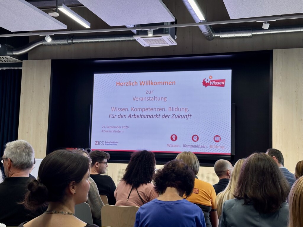 Publikum vor der Leinwand mit der Begrüßung „Herzlich Willkommen“ zur Veranstaltung „Wissen. Kompetenzen. Bildung. Für den Arbeitsmarkt der Zukunft“
